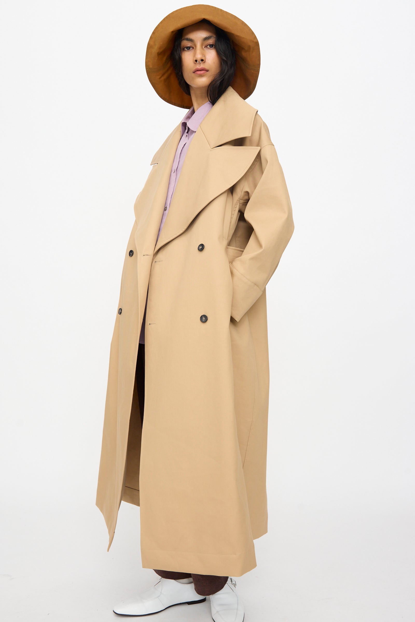 A person wears the Cawley British Cotton Gabardine Agnes Mac in Nutmeg, a wide-brimmed tan hat, a lavender shirt, brown pants, and white shoes while standing against a plain white background.