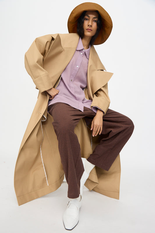 A person poses against a plain background wearing a wide-brimmed tan hat, a lavender shirt, brown pants, white shoes, and the Cawley British Cotton Garbardine Agnes Mac in Nutmeg.