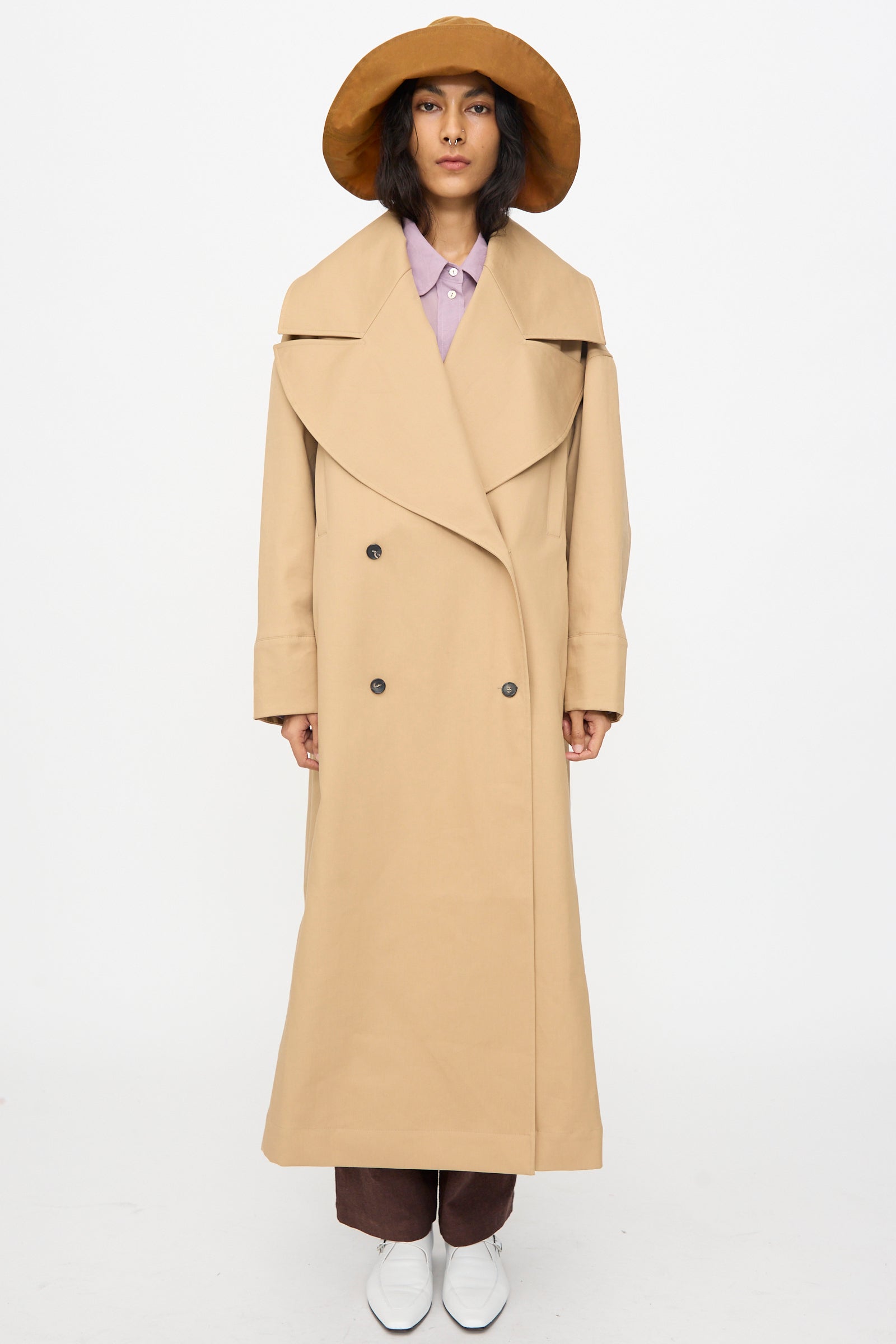 A person stands against a plain white background wearing Cawley’s British Cotton Gabardine Agnes Mac in Nutmeg, paired with a wide-brimmed tan hat, light purple shirt, brown pants, and white shoes.