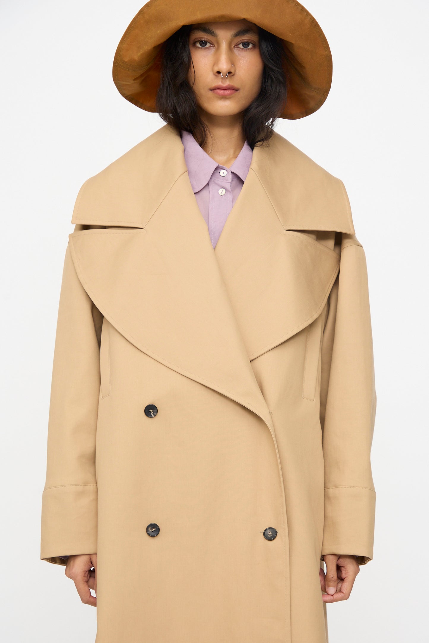 A person wearing the Cawley British Cotton Gabardine Agnes Mac in Nutmeg over a lavender collared shirt and a wide-brimmed brown hat, posed against a plain white background.
