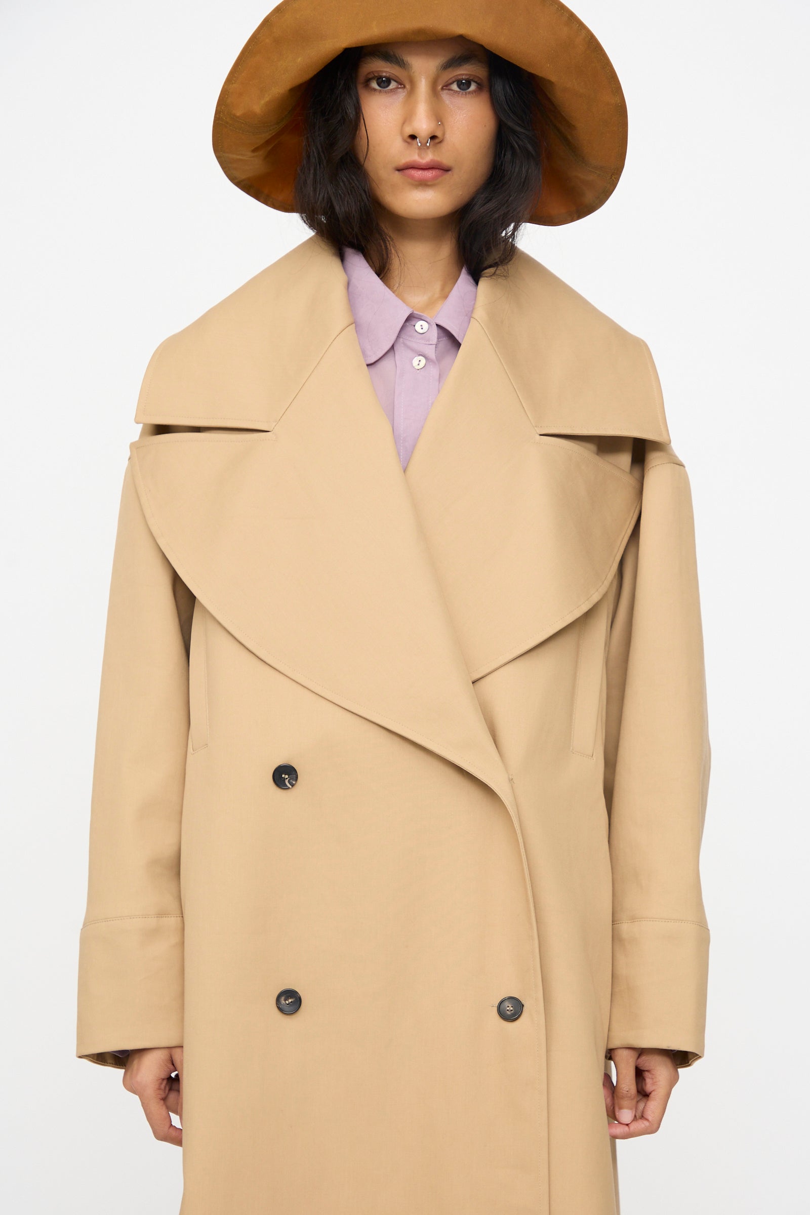 A person wearing the Cawley British Cotton Gabardine Agnes Mac in Nutmeg over a lavender collared shirt and a wide-brimmed brown hat, posed against a plain white background.