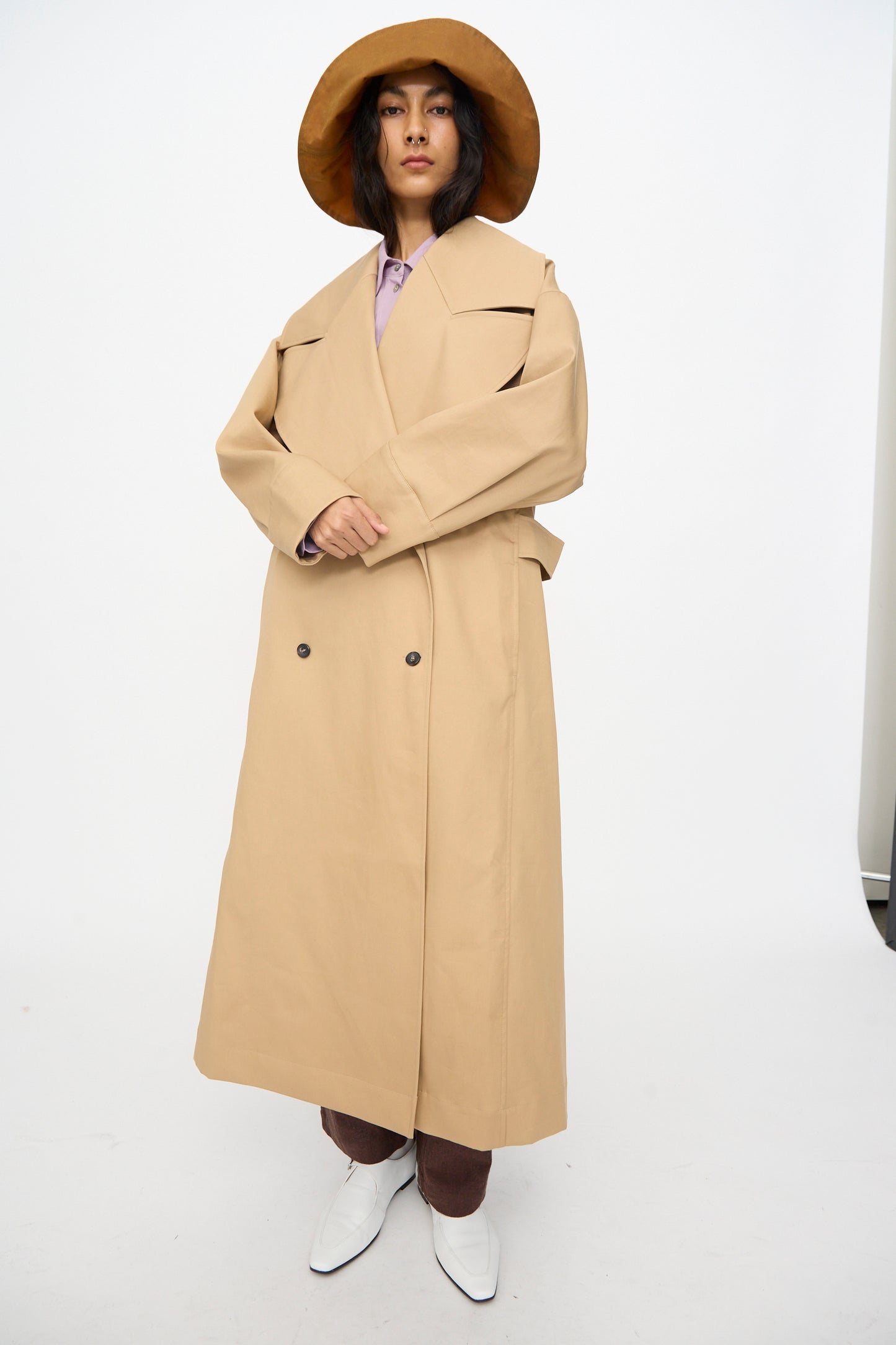 A person wearing the Cawley British Cotton Gabardine Agnes Mac in Nutmeg stands against a plain white background, styled with a wide-brimmed brown hat, brown pants, and white pointed shoes.