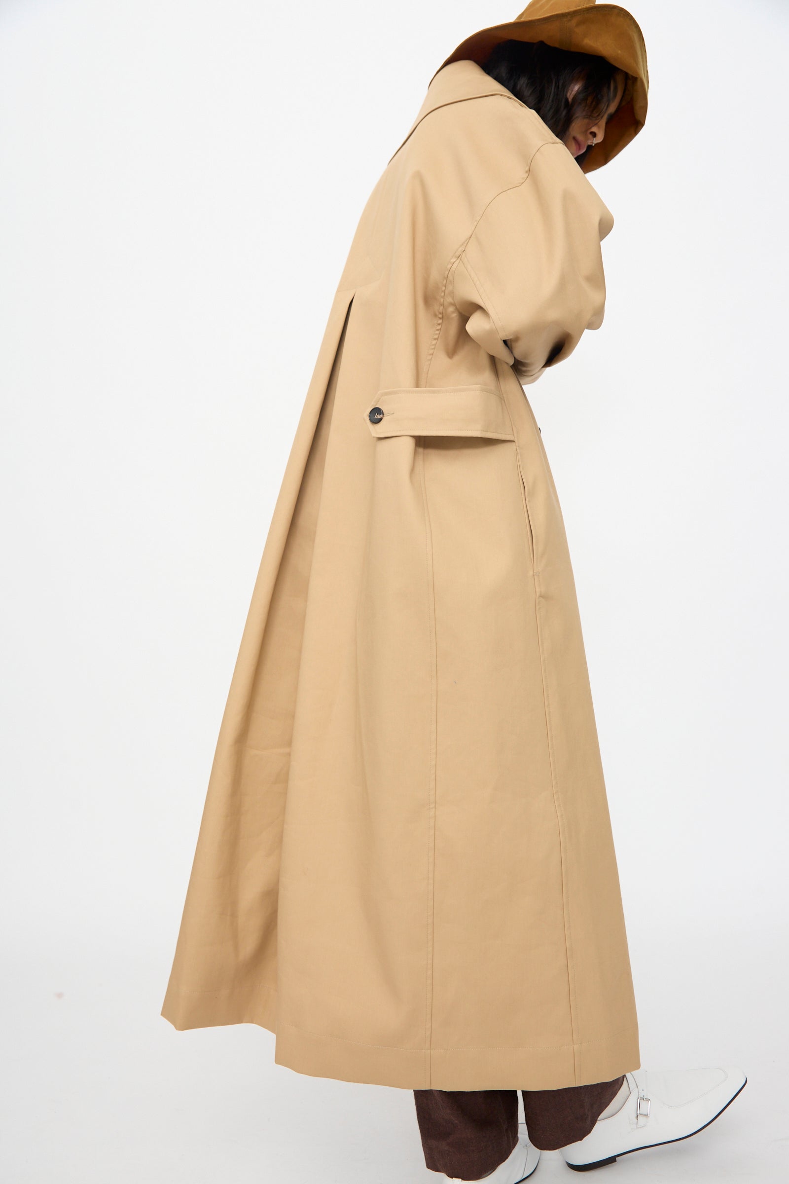 A person wears the Cawley British Cotton Gabardine Agnes Mac in Nutmeg with brown pants and white boots, standing against a plain white background.
