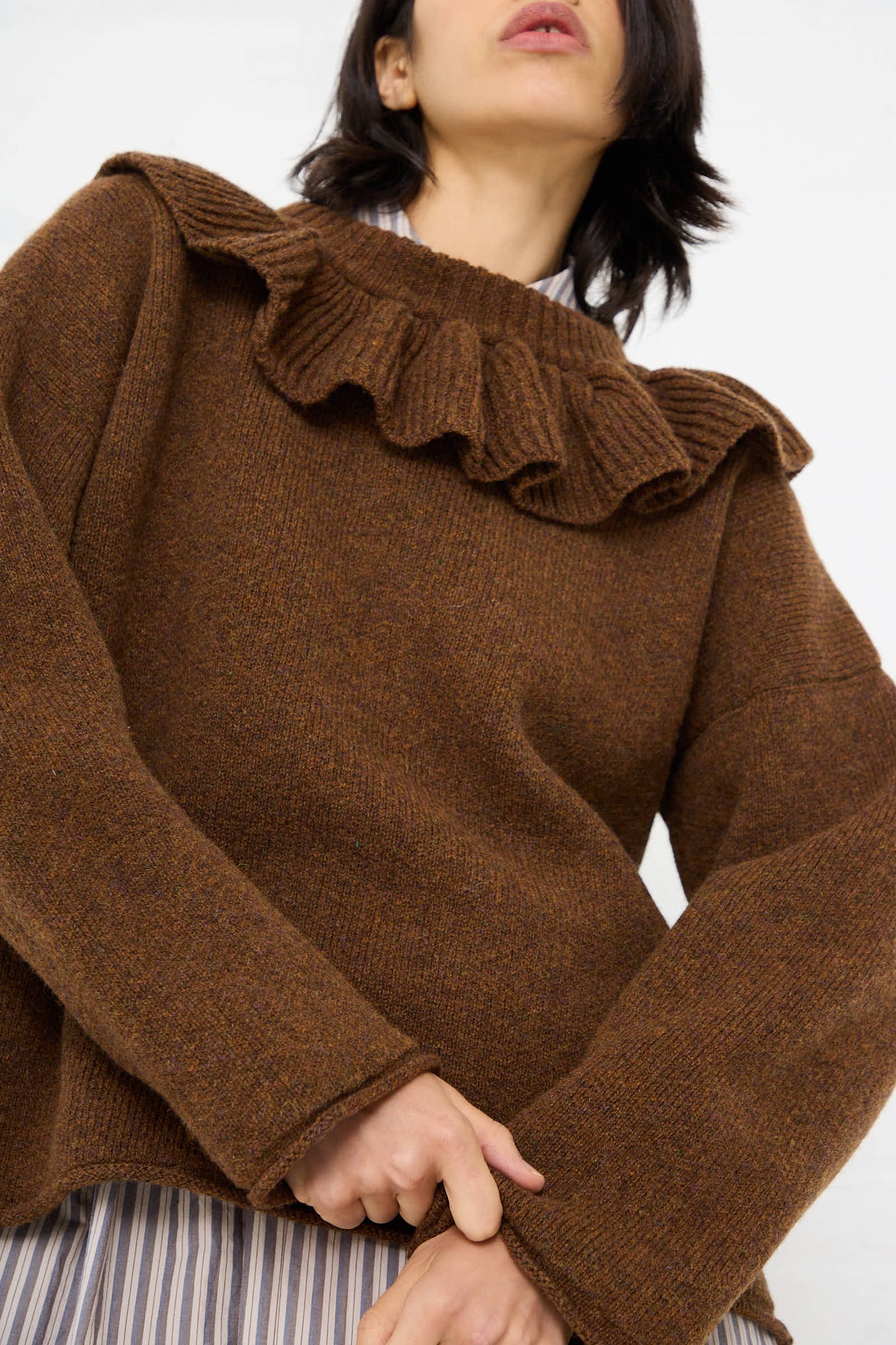 A person wearing the Cawley British Lambswool Ruffle Jumper in Tobacco, with ruffle detailing at the neckline and wide sleeves, tugging at the garment near the hem. The background is plain white.