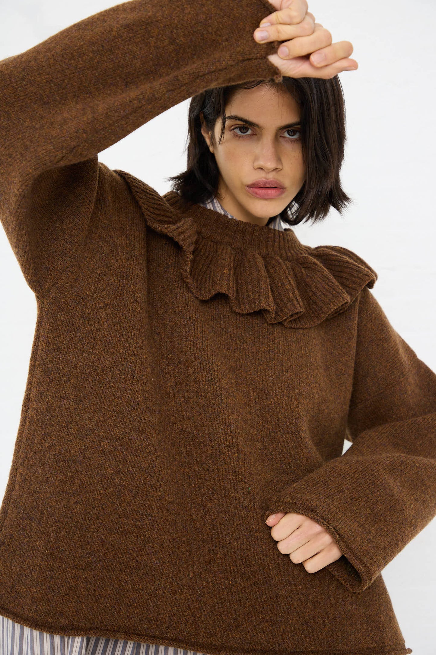 A person with short dark hair, wearing the British Lambswool Ruffle Jumper in Tobacco by Cawley, poses with one arm raised.