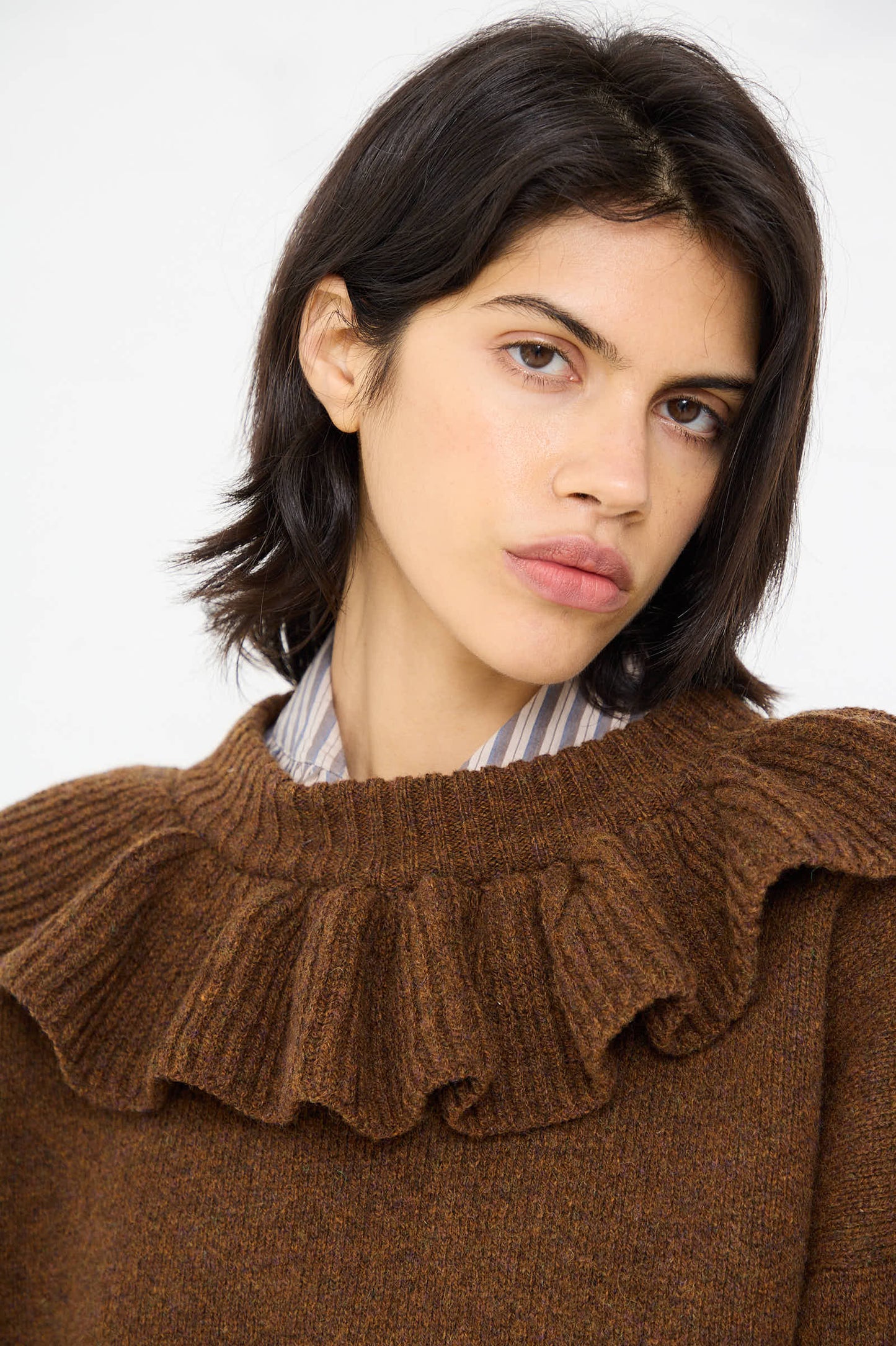 A person with shoulder-length dark hair is wearing the Cawley British Lambswool Ruffle Jumper in Tobacco over a striped shirt, looking directly at the camera.