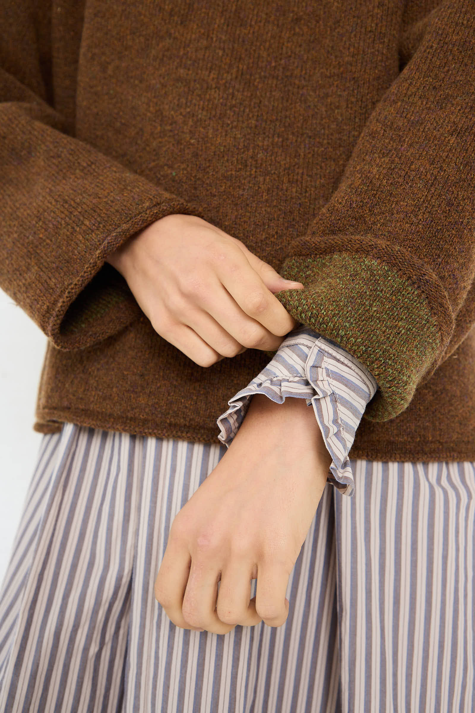 A person adjusts the sleeve of a Cawley British Lambswool Ruffle Jumper in Tobacco, revealing a striped cuff of an underlying shirt.