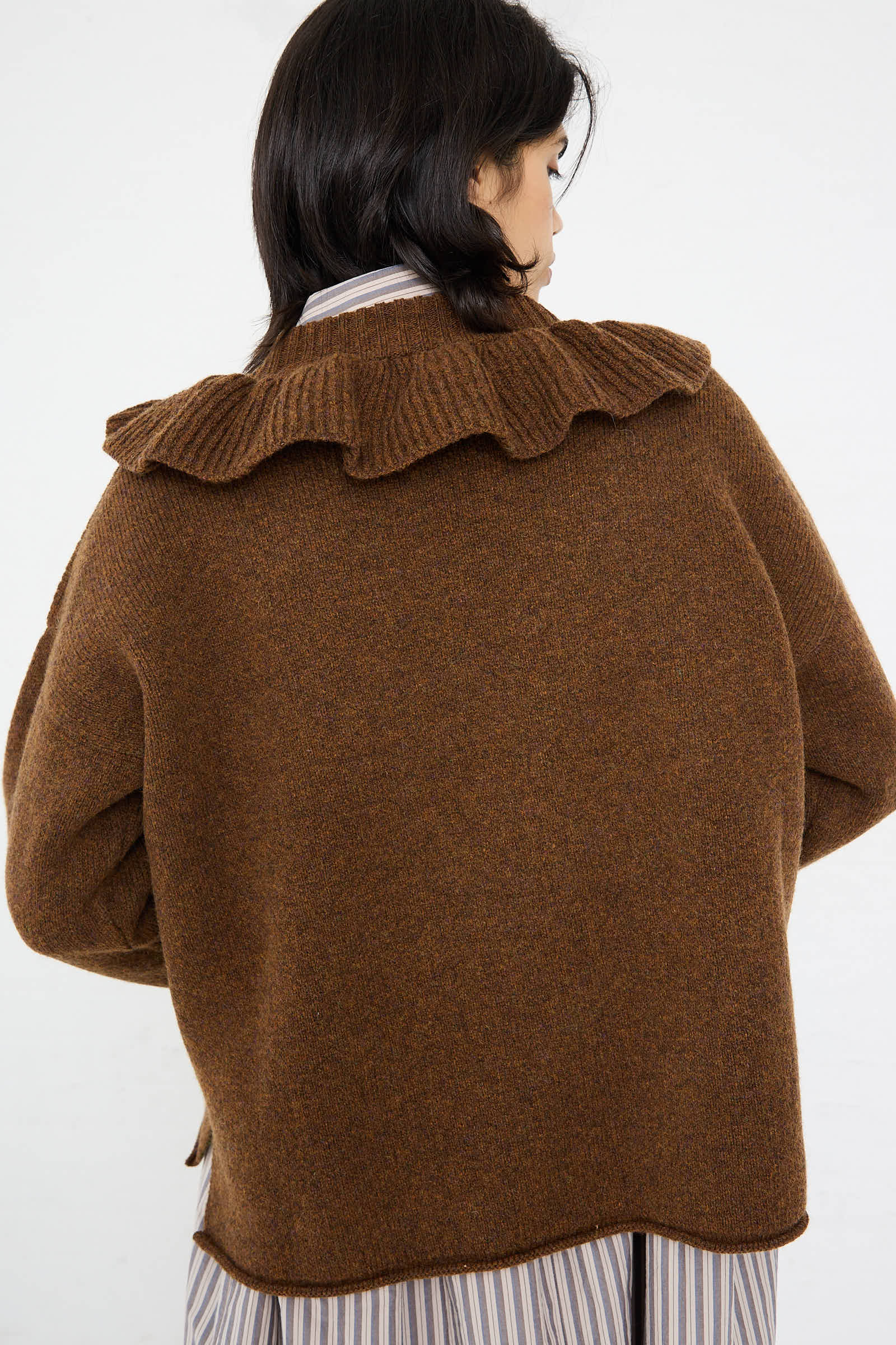 A person with dark hair facing away, wearing the Cawley British Lambswool Ruffle Jumper in Tobacco over a striped shirt.