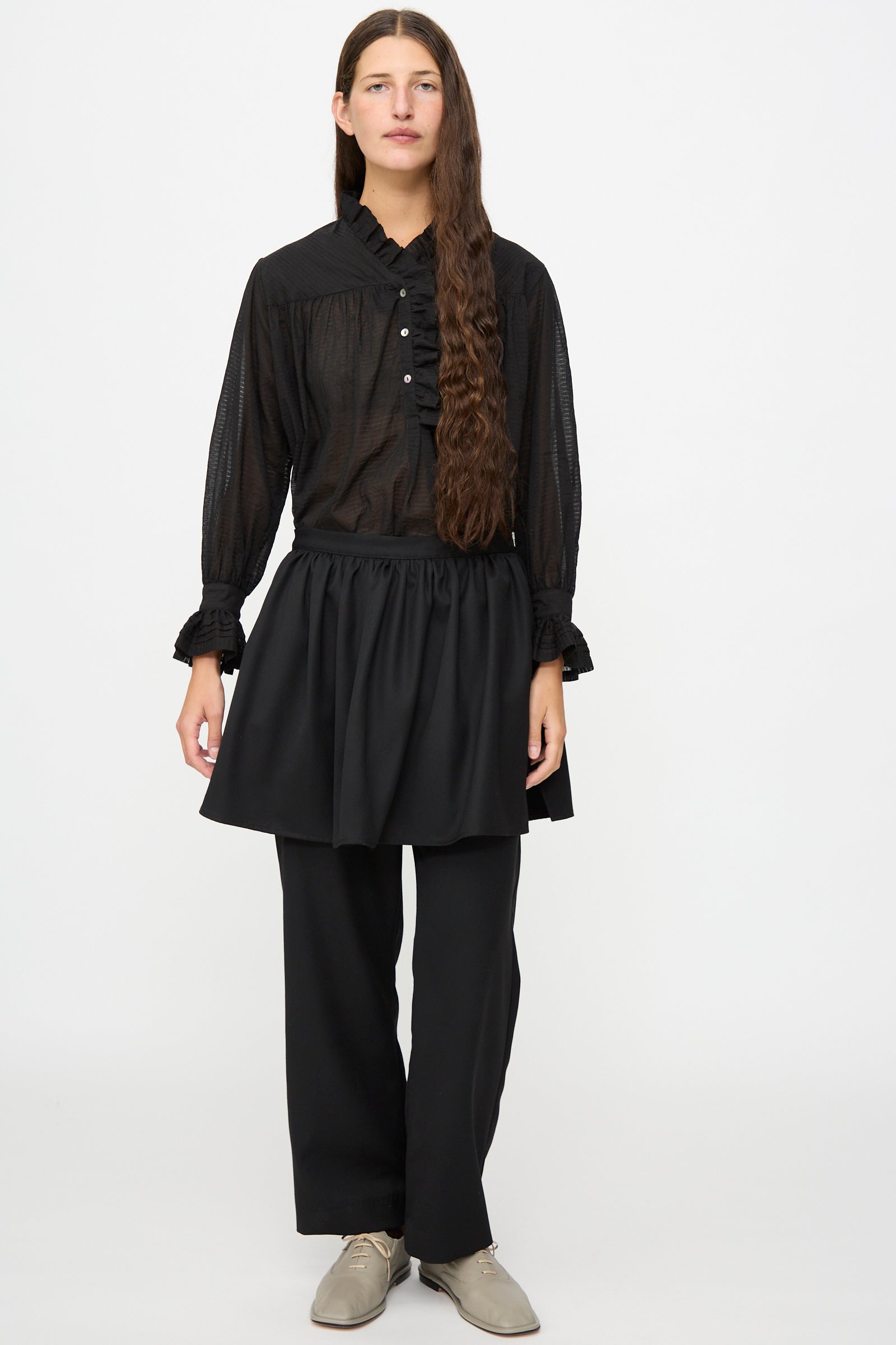A person stands against a white background wearing a black long-sleeve blouse, Cawley’s British Lightweight Wool Layered Skirt Trouser in Black, and beige shoes.