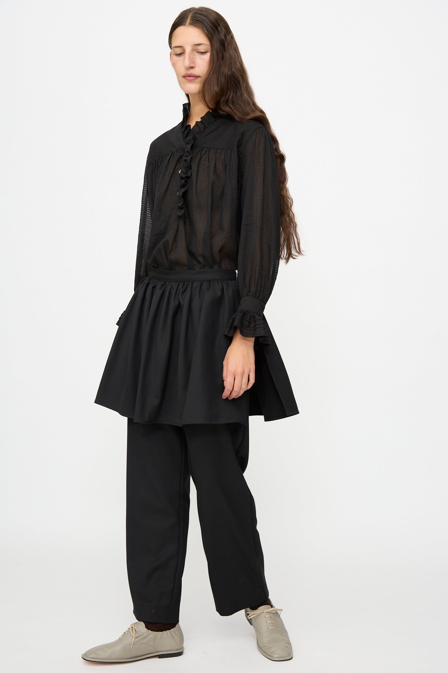 A person with long brown hair wears a sheer black blouse, the Cawley British Lightweight Wool Layered Skirt Trouser in Black, and beige shoes, standing against a plain white background.
