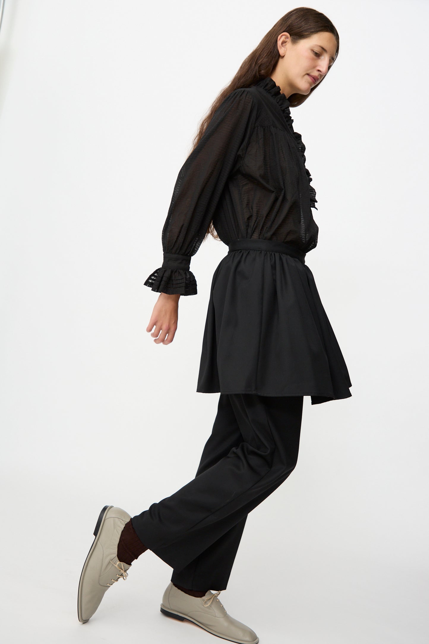 A person wearing a black ruffled blouse, Cawley’s British Lightweight Wool Layered Skirt Trouser in Black, and beige shoes walks against a plain white background.