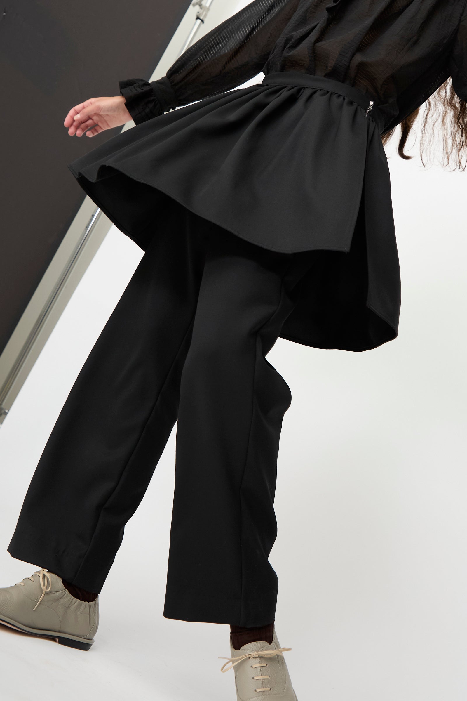 A person wears the Cawley British Lightweight Wool Layered Skirt Trouser in Black, made in the UK, styled with beige shoes and set against a plain background.
