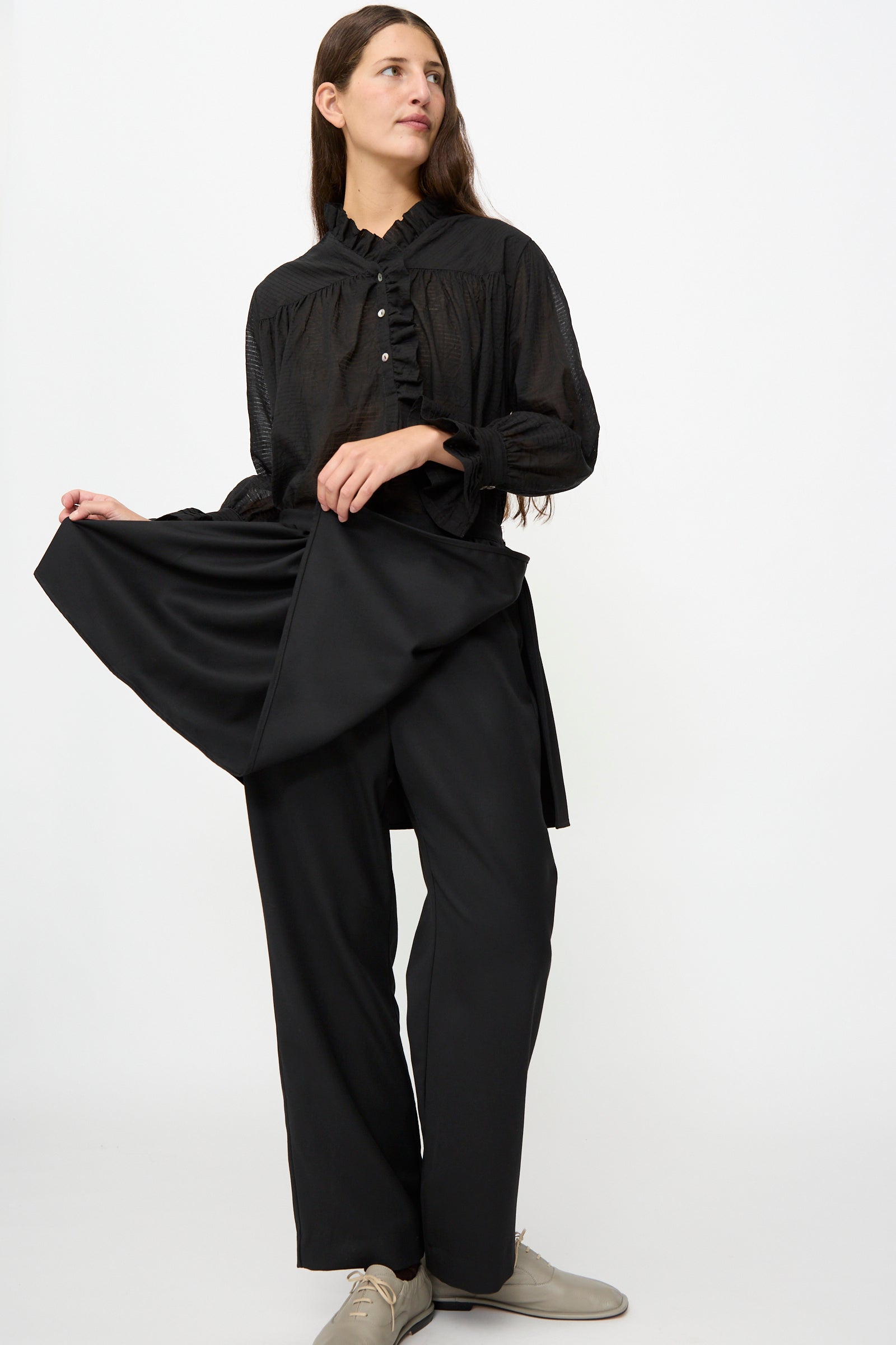 A person wearing Cawley’s British Lightweight Wool Layered Skirt Trouser in Black stands against a white background, holding out part of their outfit to the side to showcase the unique layered design crafted from lightweight wool.