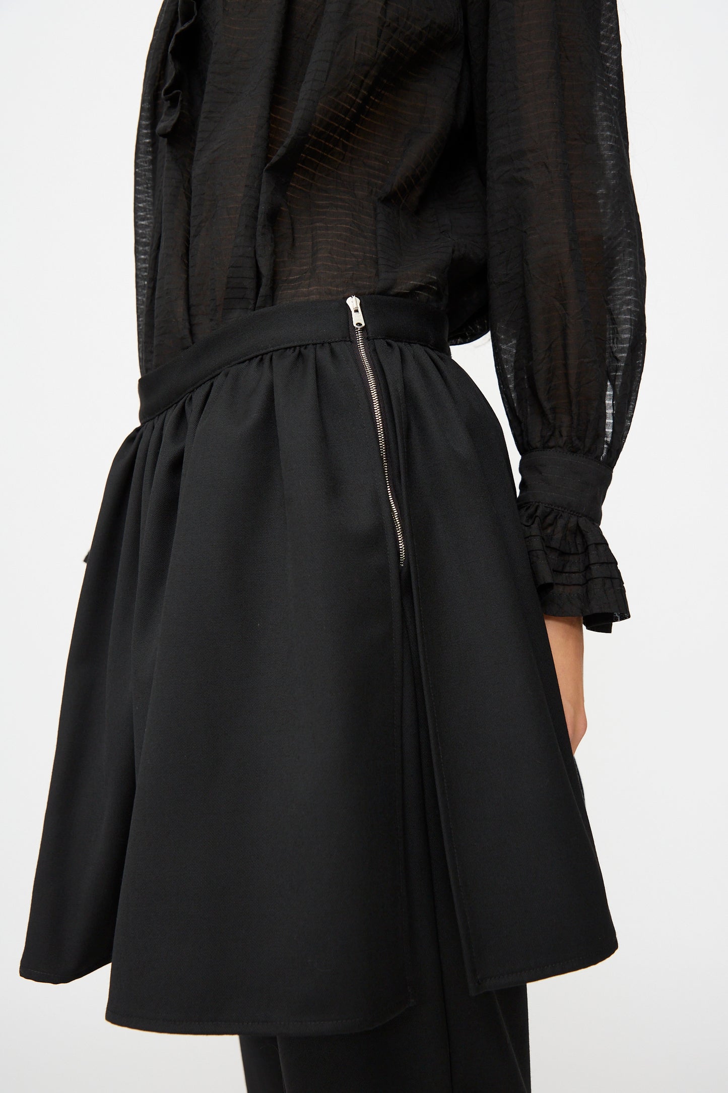 A person wearing a black textured blouse with ruffled cuffs paired with the British Lightweight Wool Layered Skirt Trouser in Black by Cawley, made in the UK, featuring a visible zipper detail on the side.