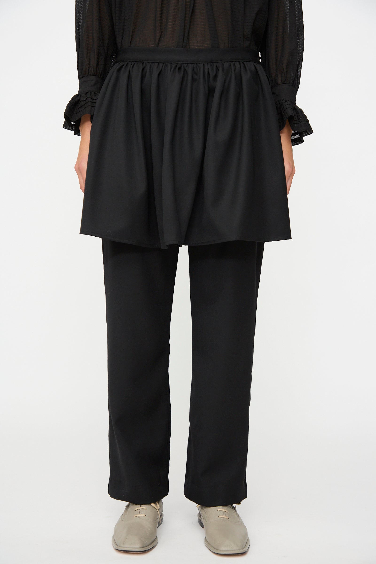 A person wearing a black sheer long-sleeve top, the Cawley British Lightweight Wool Layered Skirt Trouser in Black, and beige shoes, standing against a white background.