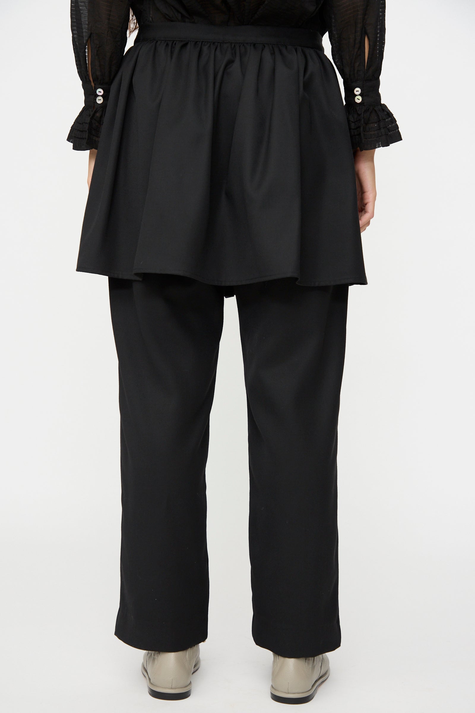A person is shown from the back wearing the Cawley British Lightweight Wool Layered Skirt Trouser in Black—black pants with an attached layered skirt overlay, crafted from lightweight wool and made in the UK. The outfit is styled with a black long-sleeve shirt featuring buttoned cuffs and paired with light-colored shoes.