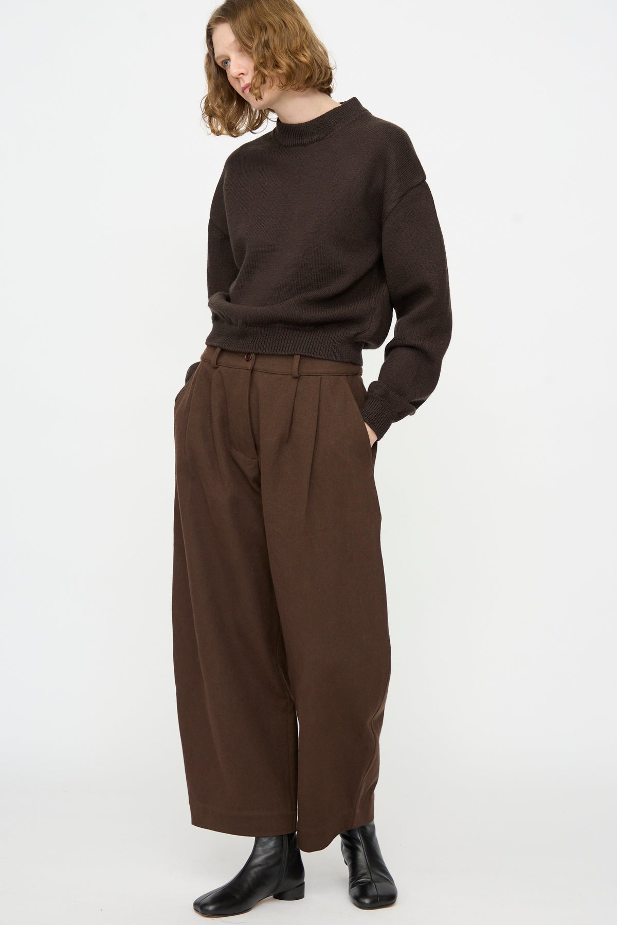 A person stands against a plain background, wearing the Cawley British Merino Cindy Crewneck Jumper in Bark, paired with wide-legged brown pants and black ankle boots, hands in pockets and head turned slightly to the side.