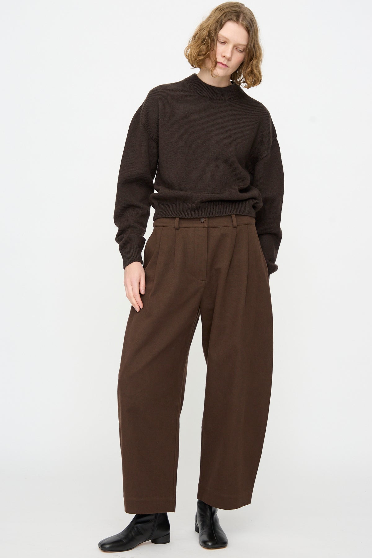 A person wearing the Cawley British Merino Cindy Crewneck Jumper in Bark, paired with loose brown pleated pants and black ankle boots, stands against a plain white background.