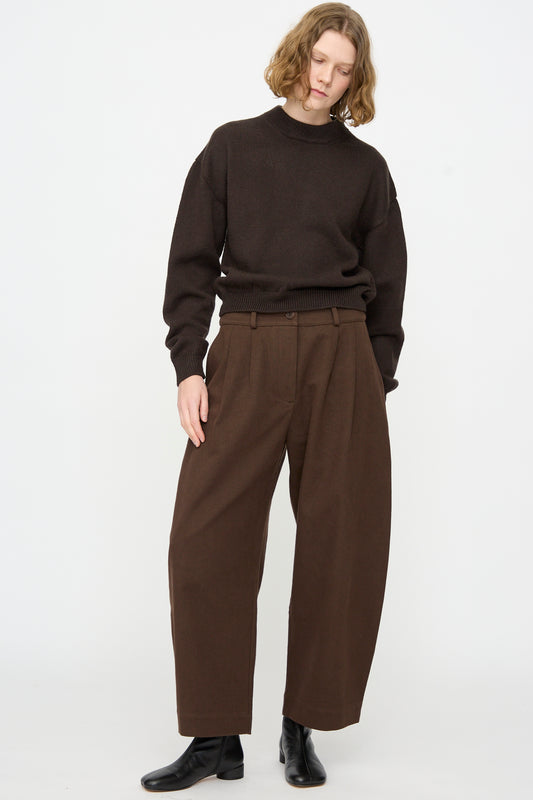 A person wearing the Cawley British Merino Cindy Crewneck Jumper in Bark, paired with loose brown pleated pants and black ankle boots, stands against a plain white background.