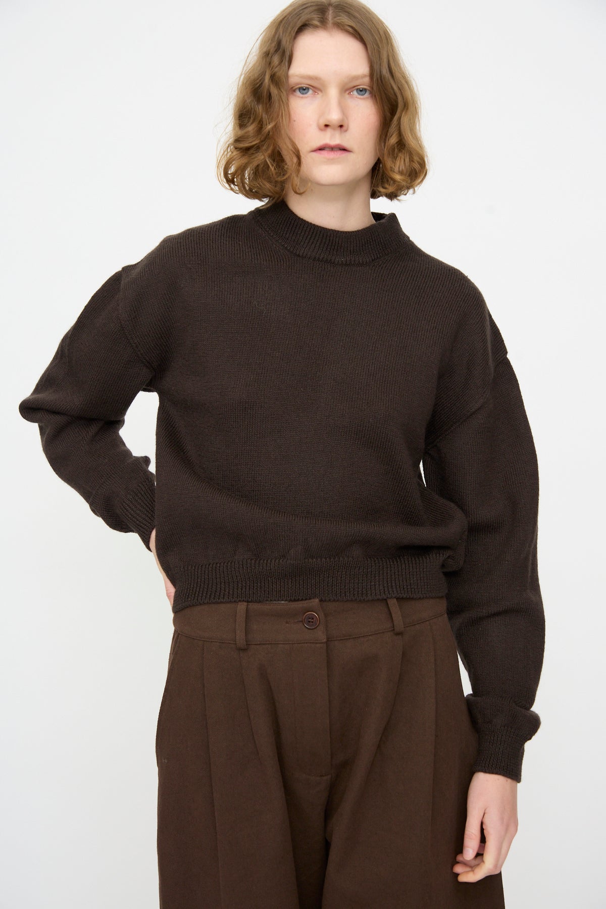 A person with chin-length wavy hair wears the Cawley British Merino Cindy Crewneck Jumper in Bark and brown trousers, standing against a plain white background.