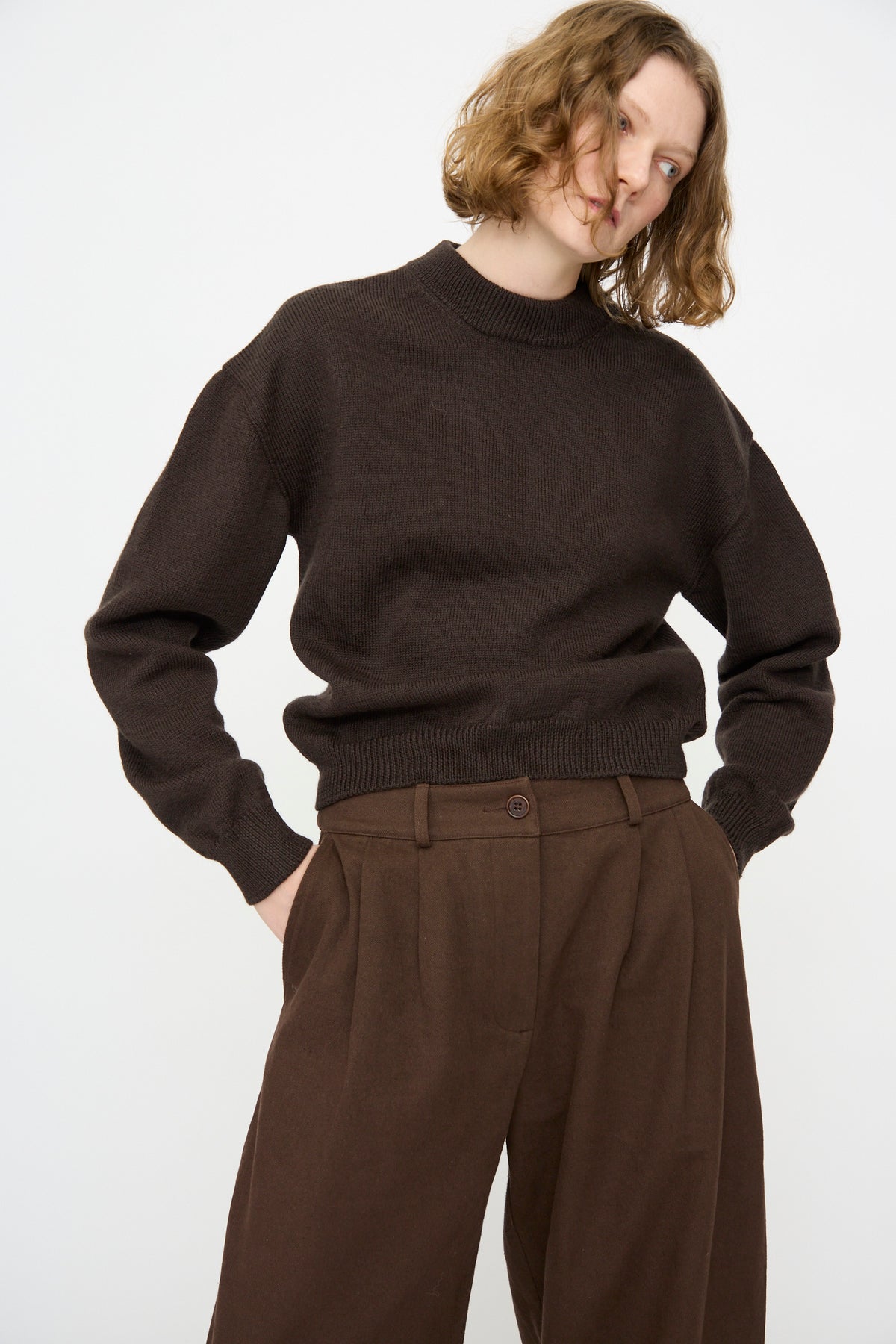 A person wearing the Cawley British Merino Cindy Crewneck Jumper in Bark and brown trousers stands against a plain white background with hands in their pockets.