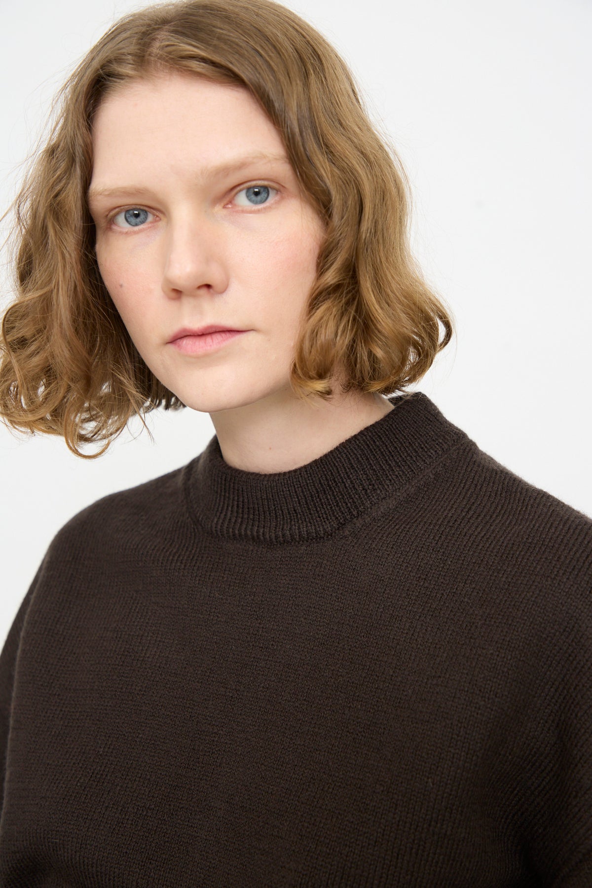 A person with short, wavy light brown hair wears the Cawley British Merino Cindy Crewneck Jumper in Bark and looks at the camera against a plain light background.