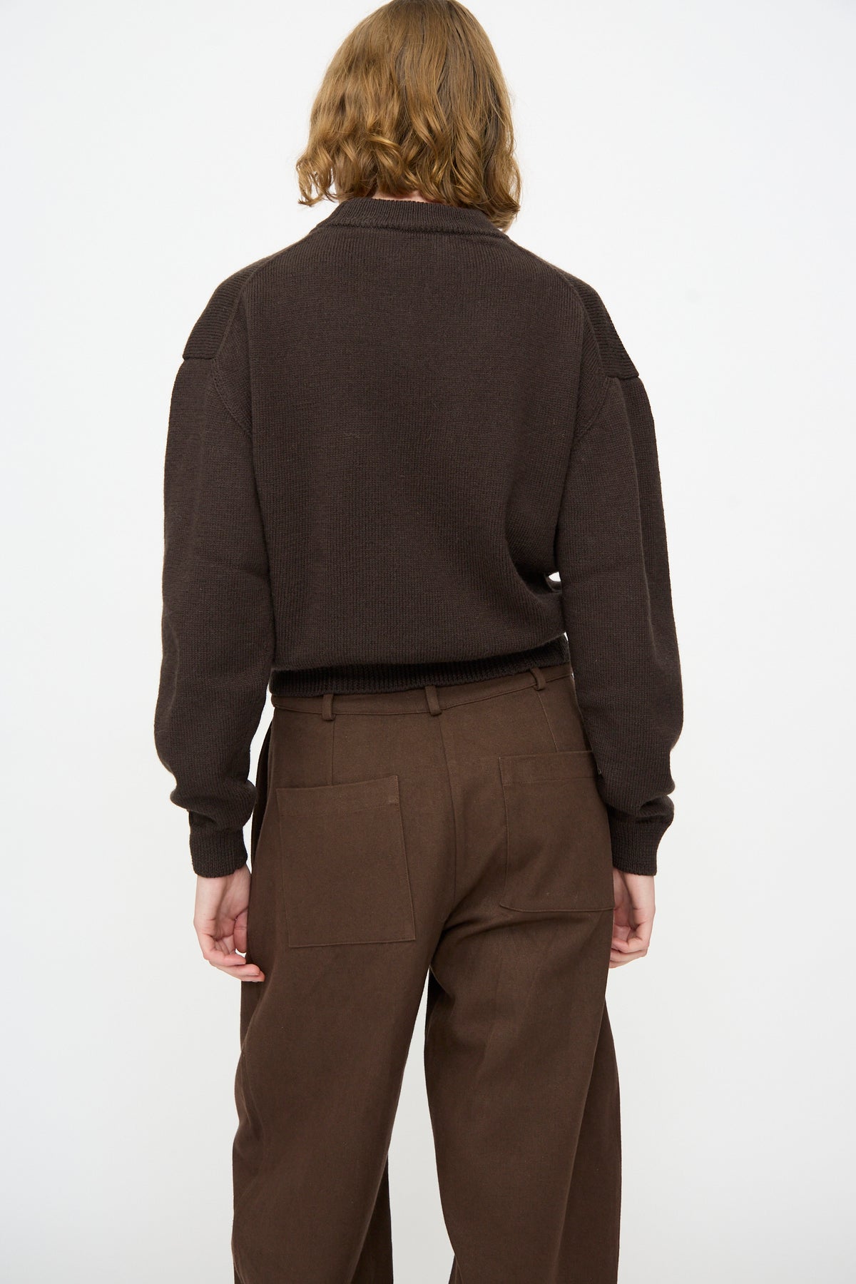 A person with shoulder-length hair is shown from the back against a plain white background wearing the Cawley British Merino Cindy Crewneck Jumper in Bark and brown pants.