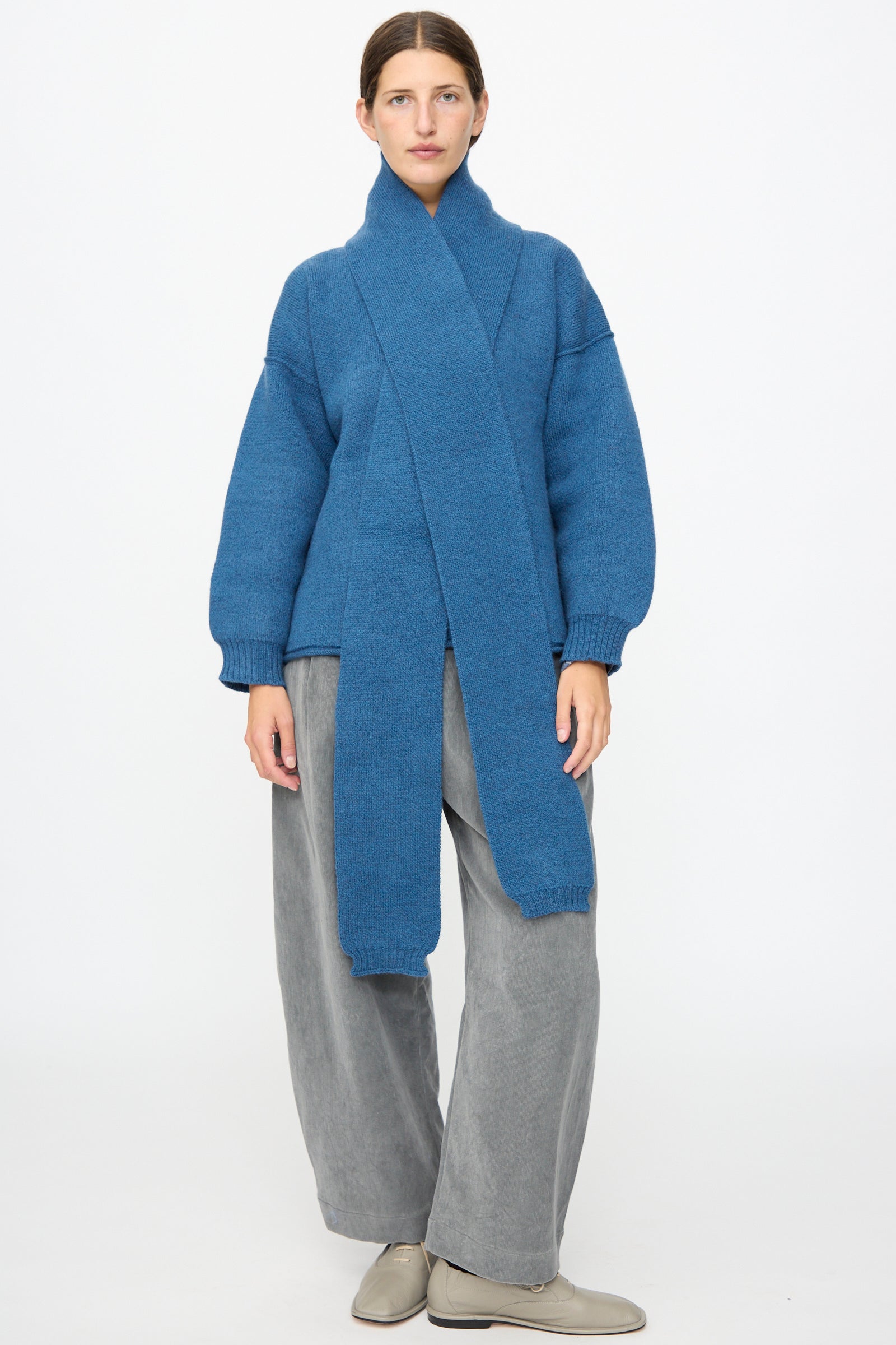 A person stands facing forward against a plain white background, wearing the Cawley British Merino Wool Long Scarf in Sea—an attached long knit scarf crafted from soft merino wool—styled with a blue sweater, loose gray pants, and beige shoes.