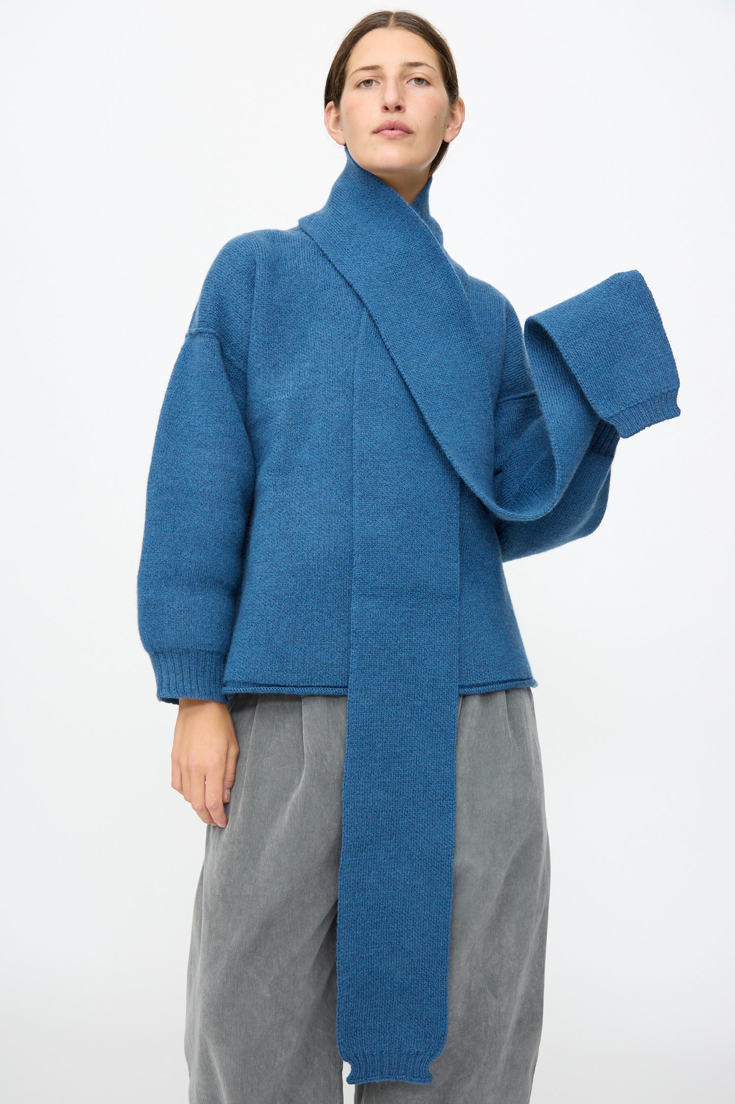 A person wears the Cawley British Merino Wool Long Scarf in Sea, paired with a blue sweater and loose grey pants, standing against a plain white background.