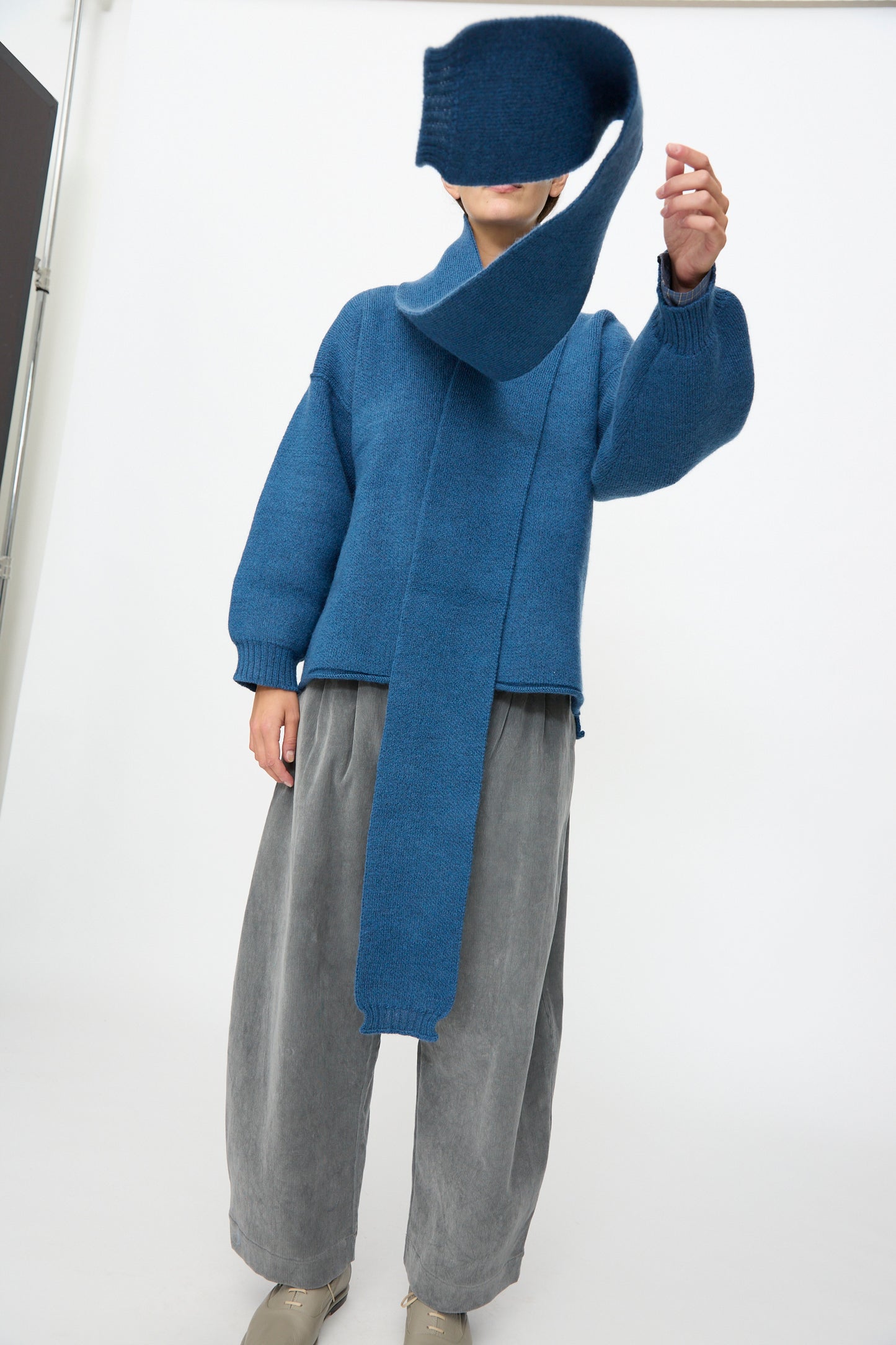 A person is seen wearing a blue sweater and the Cawley British Merino Wool Long Scarf in Sea, paired with wide gray pants and beige shoes, holding the end of the scarf in front of their face against a plain background.