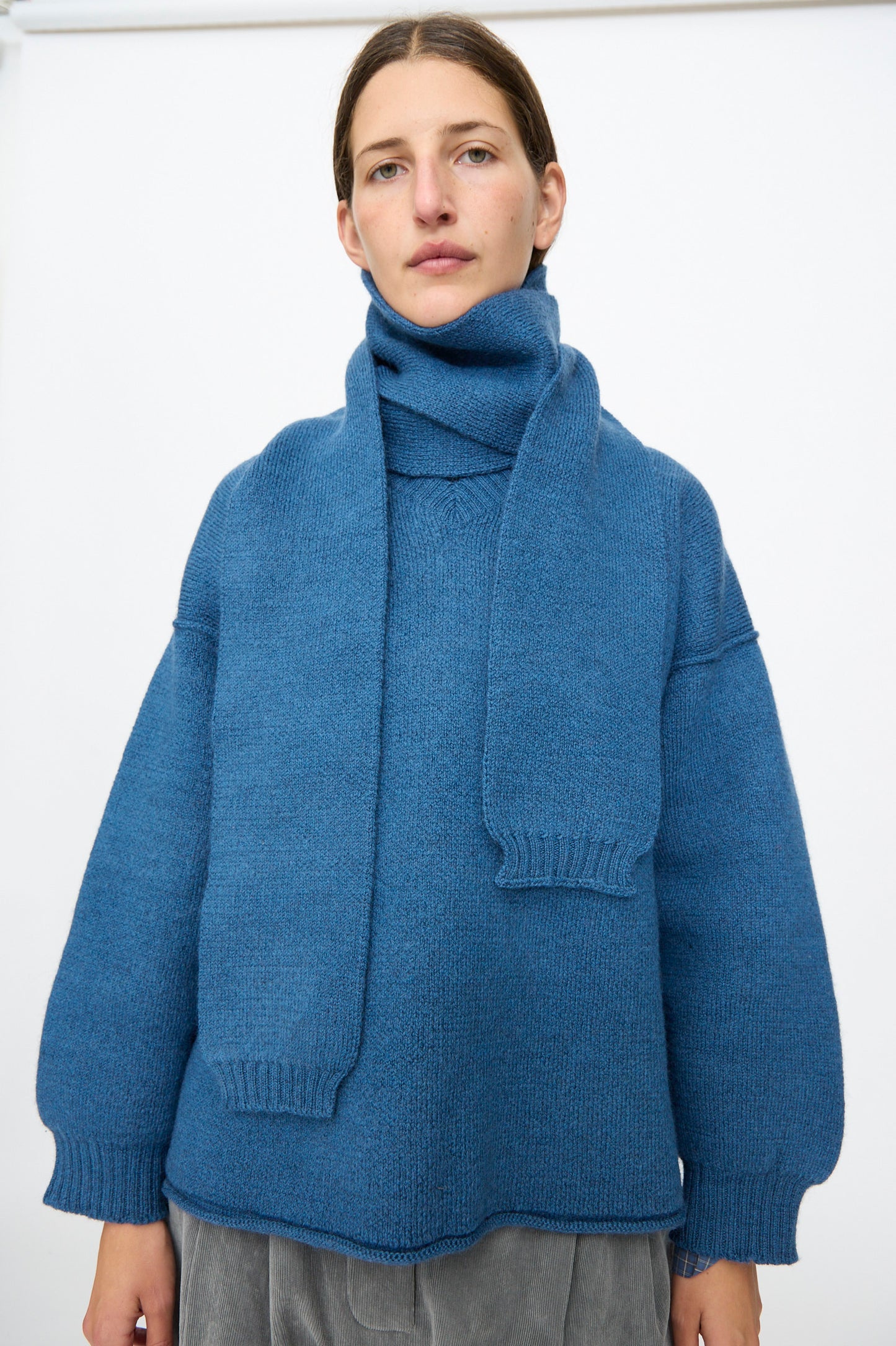 A person wearing a blue knit sweater and the Cawley British Merino Wool Long Scarf in Sea stands against a plain white background, facing the camera with a neutral expression.