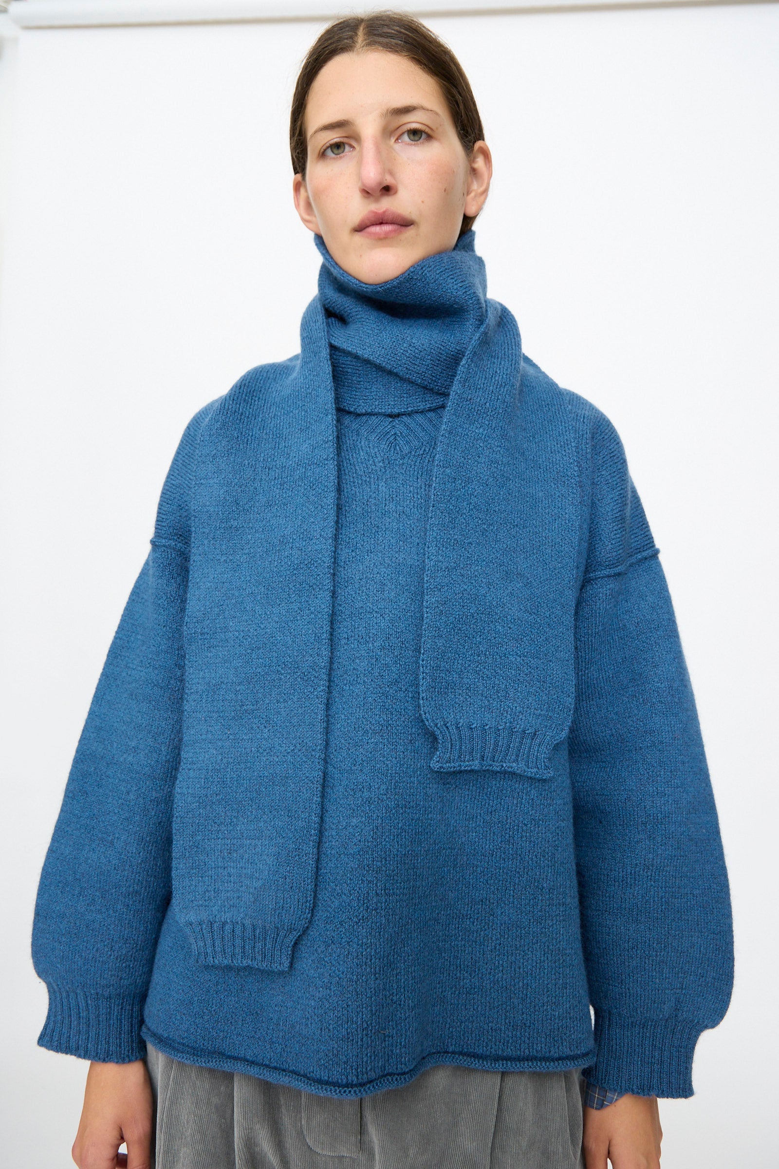 A person wearing a blue knit sweater and the Cawley British Merino Wool Long Scarf in Sea stands against a plain white background, facing the camera with a neutral expression.