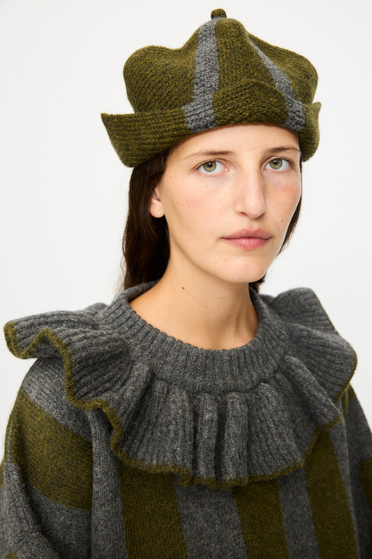 A person wears the Cawley British Merino Wool Ski Hat in Moss and Slate, paired with a matching wide-striped ruffled sweater, looking at the camera with a neutral expression.