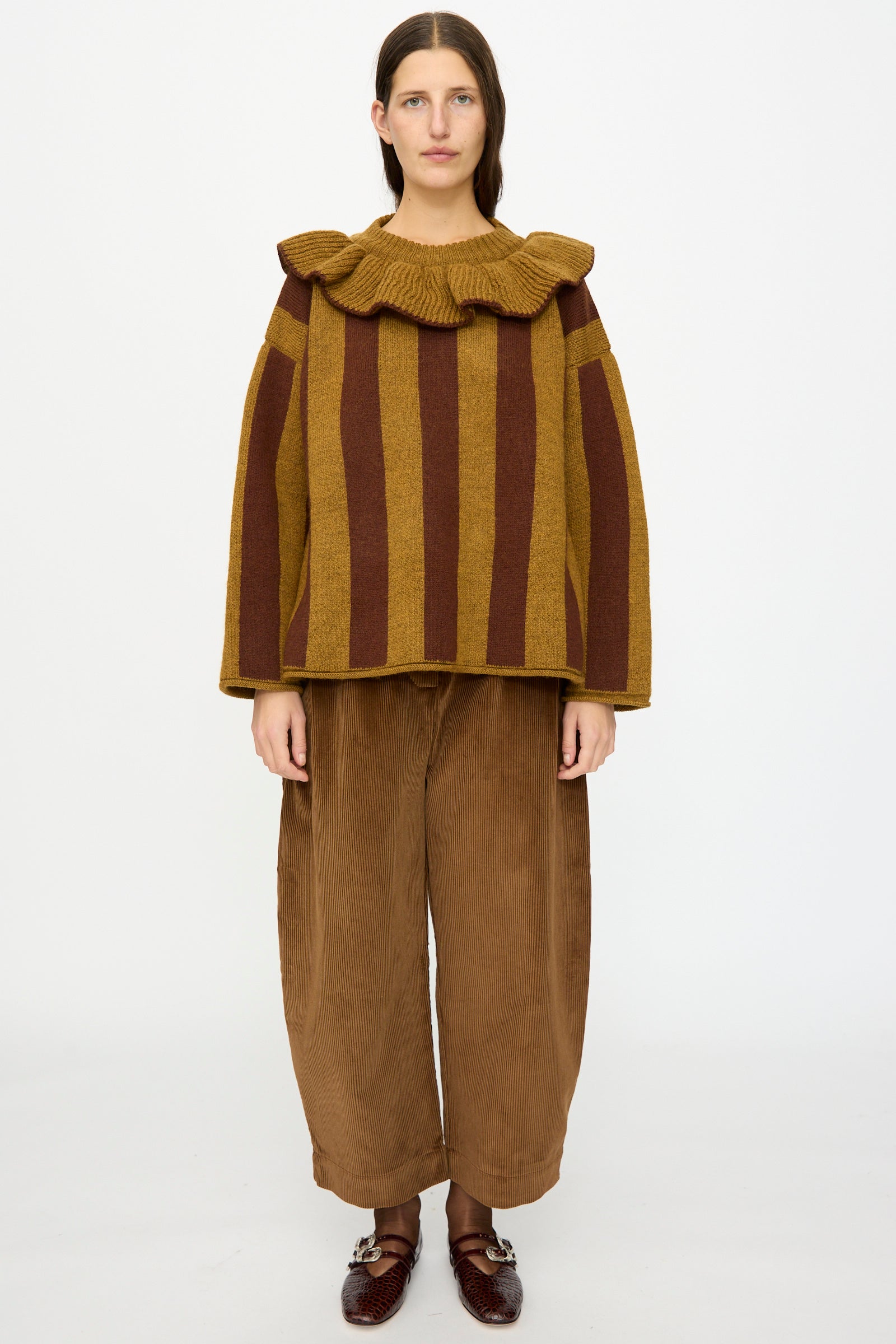 A person stands against a plain background, wearing the Cawley British Merino Wool Striped Ruffle Jumper in Goldfinch and Bordeaux, paired with wide brown pants and dark brown shoes.