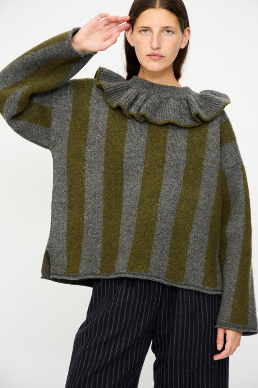 A woman wears the Cawley British Merino Wool Striped Ruffle Jumper in Slate and Moss with dark pinstriped pants, standing against a plain background.