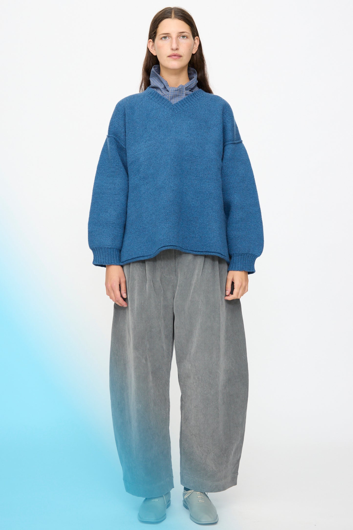 A person stands against a plain background, wearing the Cawley British Merino Wool V-Neck Jumper in Sea, paired with gray wide-leg pants, a blue turtleneck, and light blue shoes.