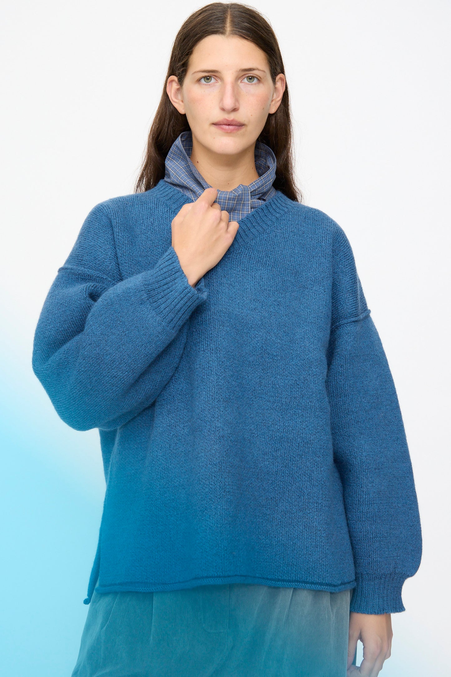 A woman with long brown hair wears the Cawley British Merino Wool V-Neck Jumper in Sea over a blue collared shirt, standing against a light gradient background and looking at the camera with a neutral expression. Crafted in the UK from fine British merino wool, this jumper blends classic style with superior craftsmanship.