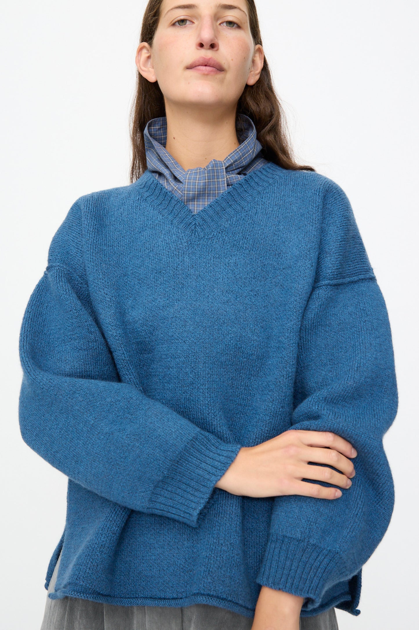 A person stands against a plain white background with their arms crossed loosely, wearing the Cawley British Merino Wool V-Neck Jumper in Sea layered over a blue plaid collared shirt.