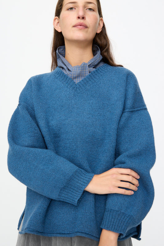 A person stands against a plain white background with their arms crossed loosely, wearing the Cawley British Merino Wool V-Neck Jumper in Sea layered over a blue plaid collared shirt.