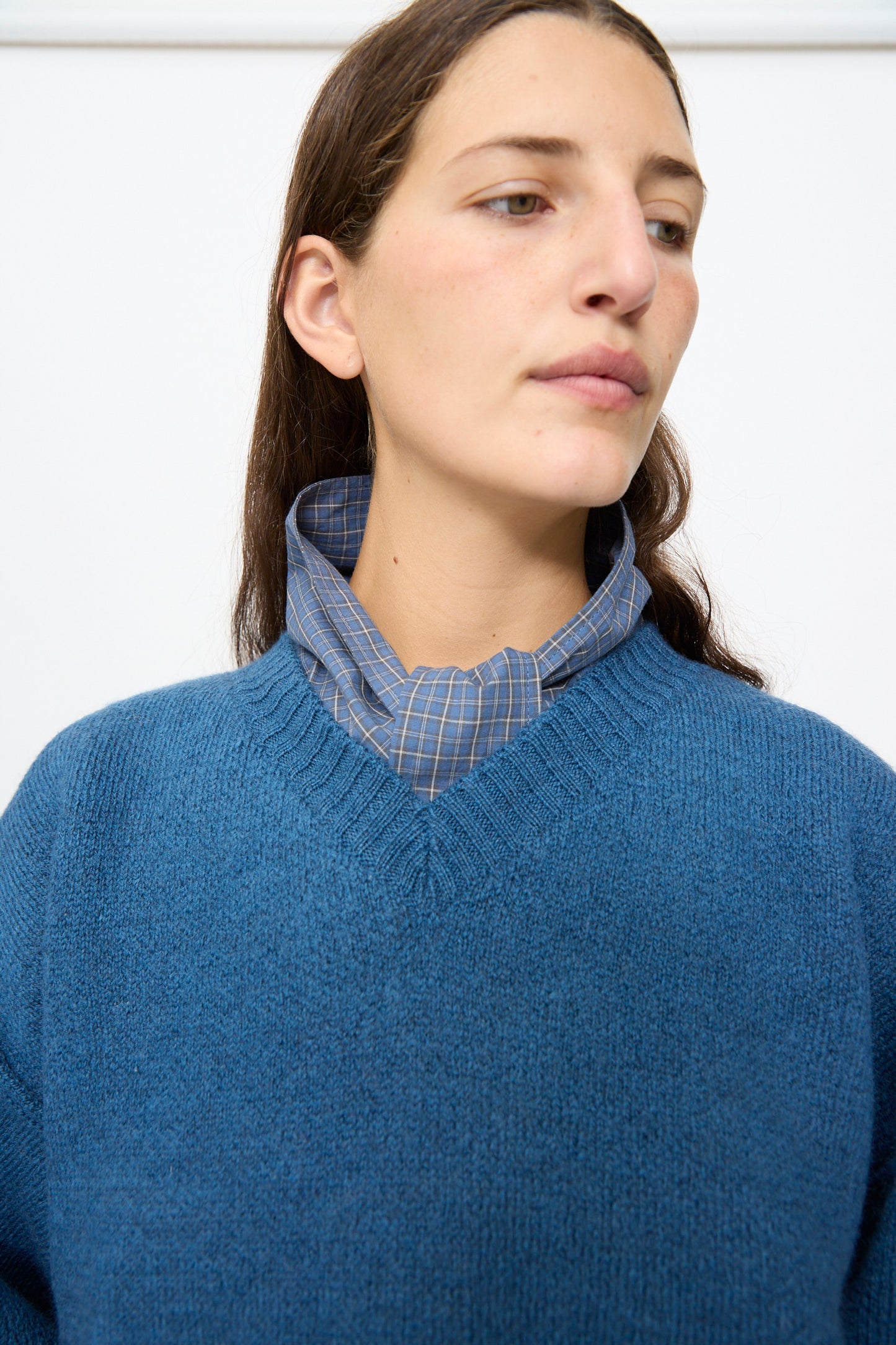 A person wearing the Cawley British Merino Wool V-Neck Jumper in Sea, made in the UK, layered over a blue plaid shirt, looks off to the side against a plain background.