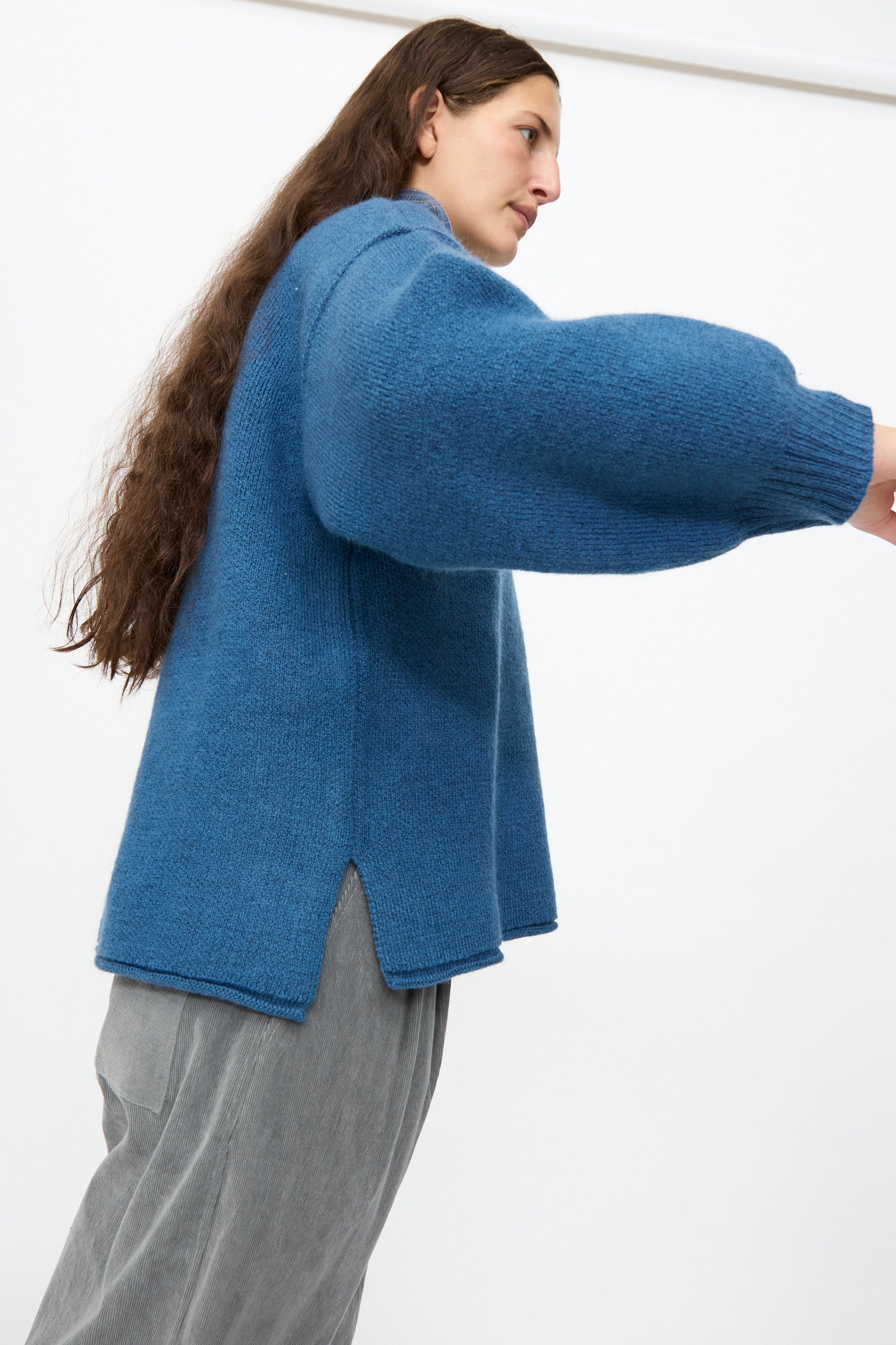 A person wearing the Cawley British Merino Wool V-Neck Jumper in Sea—an oversized blue sweater featuring side slits and wide sleeves—paired with grey pants, standing against a plain white background.