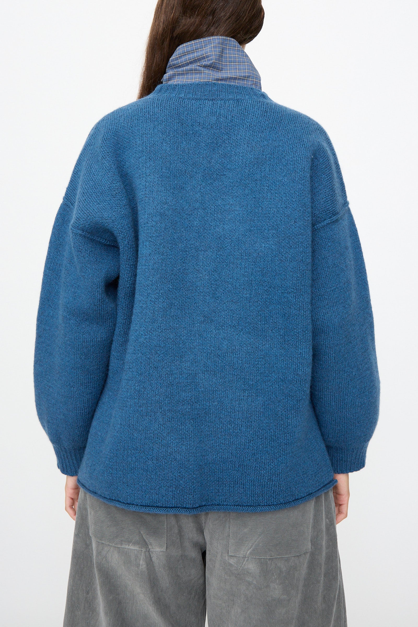 A person wears the Cawley British Merino Wool V-Neck Jumper in Sea over a collared shirt with gray pants, shown from the back against a plain white background. This sweater adds both comfort and timeless style to the look.