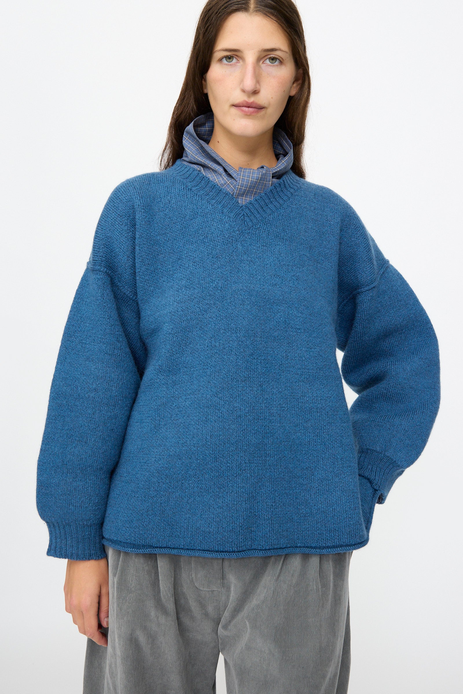 A person stands facing forward against a plain white background, wearing the Cawley British Merino Wool V-Neck Jumper in Sea over a collared shirt and gray pants.
