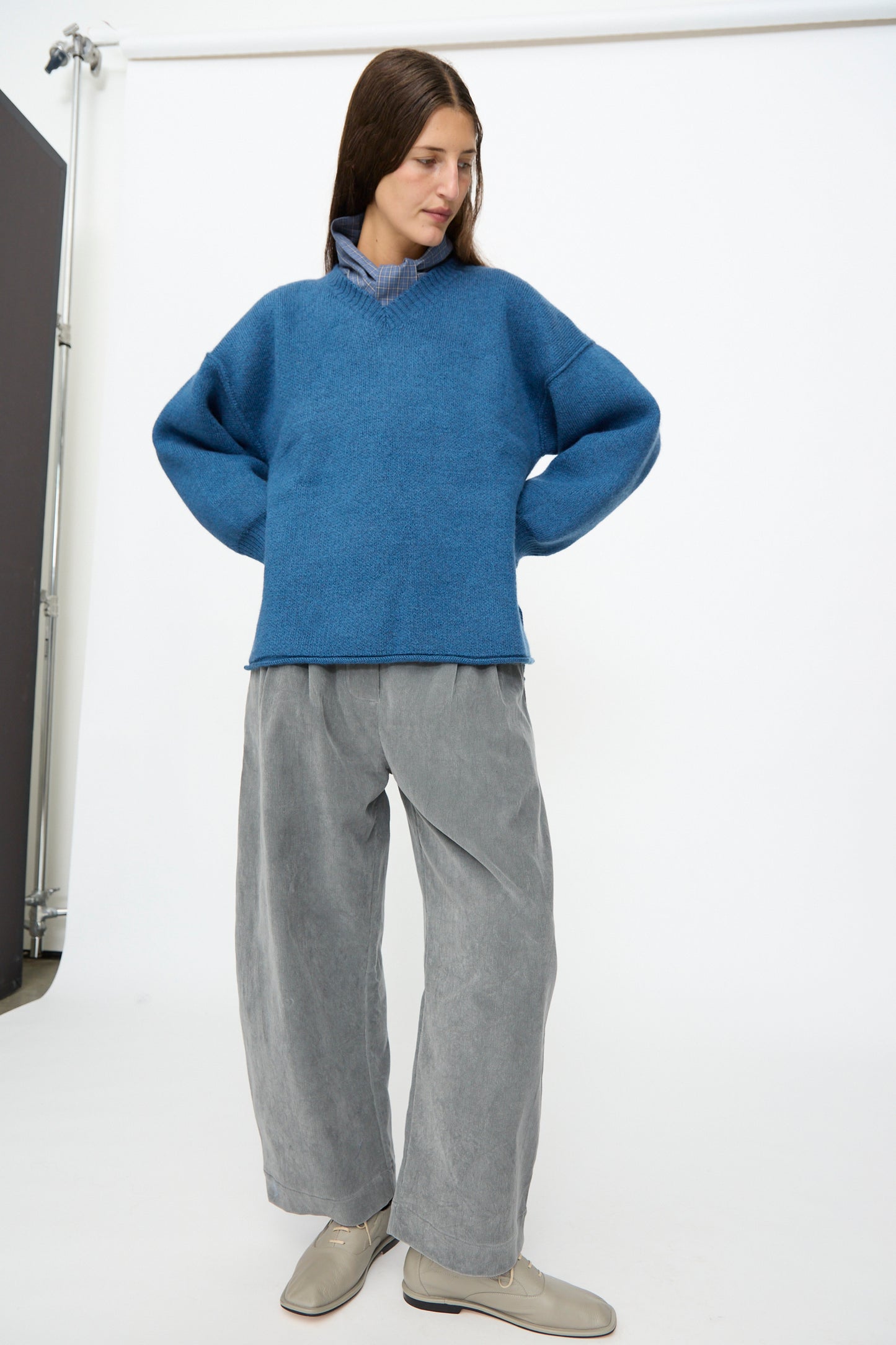 A person stands in a studio with hands on hips, looking slightly to the side, wearing the Cawley British Merino Wool V-Neck Jumper in Sea, paired with gray wide-leg pants and beige shoes.