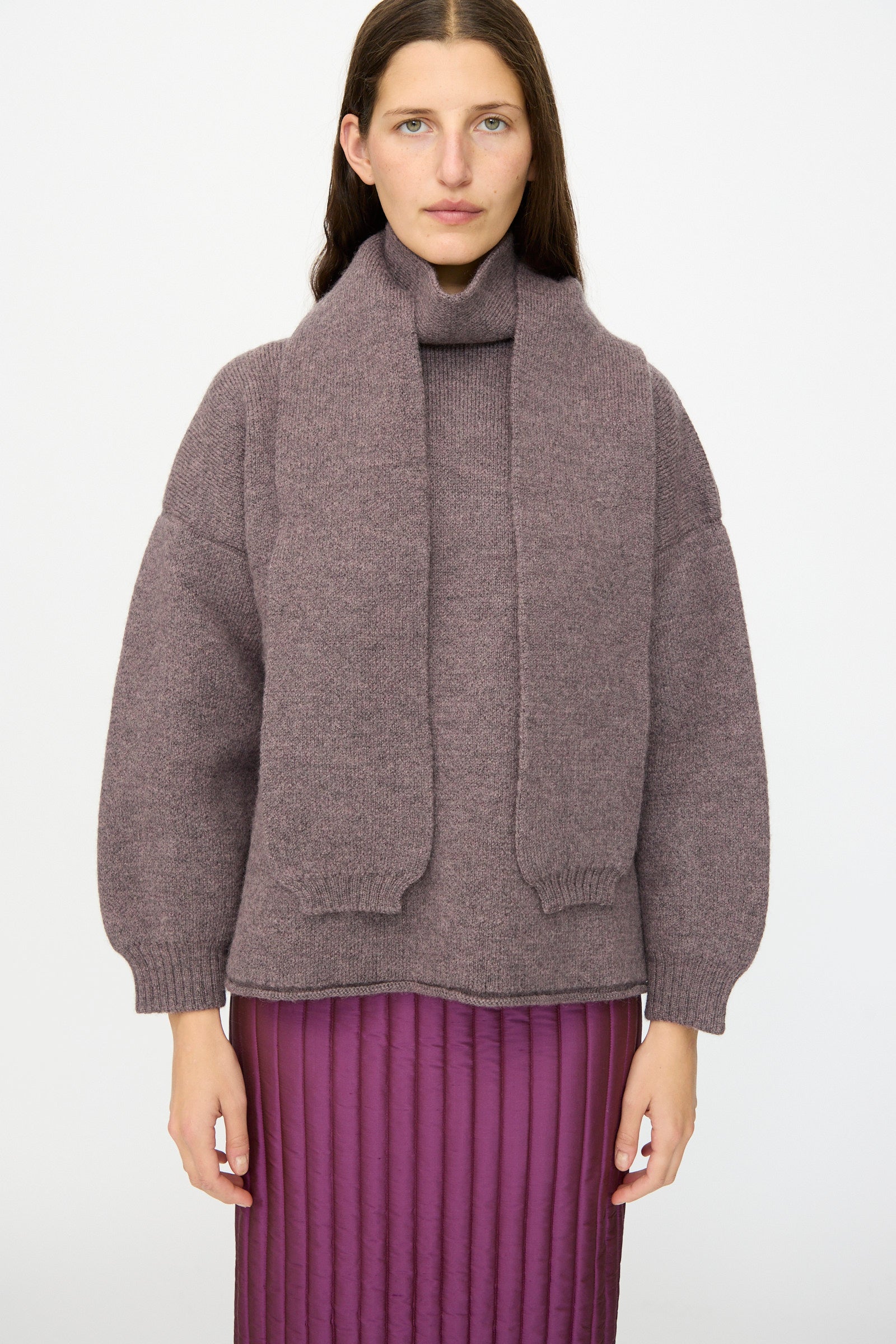 A person wearing the Cawley British Merino Wool Wrap Neck Pullover in Luna with an attached scarf and a purple quilted skirt stands against a plain white background.