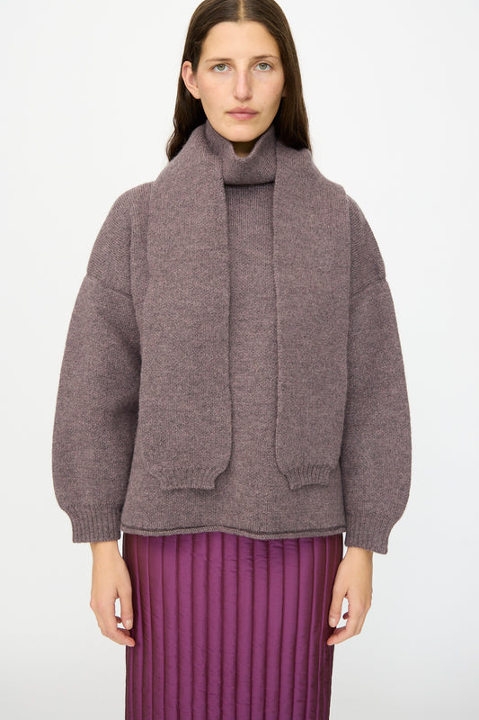 A person wearing the Cawley British Merino Wool Wrap Neck Pullover in Luna with an attached scarf and a purple quilted skirt stands against a plain white background.
