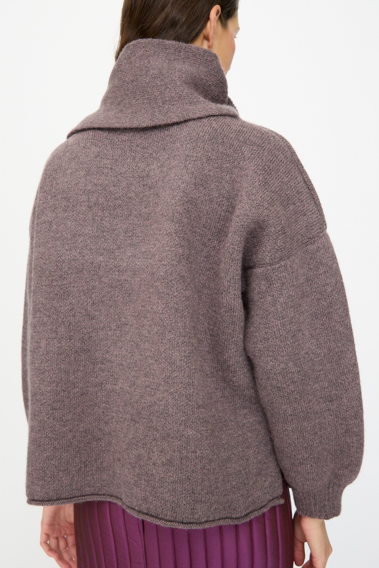 A woman is seen from the back wearing the Cawley British Merino Wool Wrap Neck Pullover in Luna, styled with a pleated purple skirt.