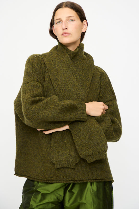 A person wears the Cawley British Merino Wool Wrap Neck Pullover in Moss, paired with a matching scarf and shiny green pants, standing against a plain white background.