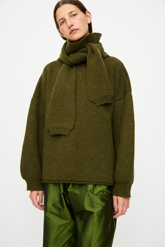 A person wearing the Cawley British Merino Wool Wrap Neck Pullover in Moss stands against a plain white background, paired with shiny green satin pants.