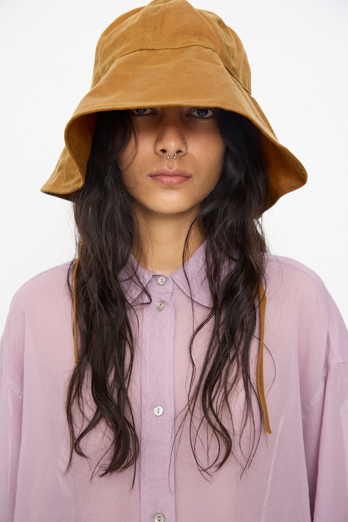 A person with long wavy hair and a nose piercing wears the Cawley British Waxed Oilskin Sunhat in Gold and a light purple button-up shirt, facing forward against a plain background.